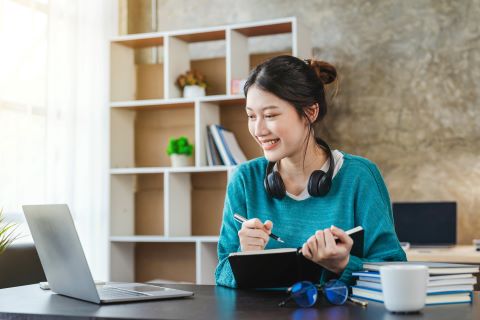 Woman sitting at a desk holding a notebook and smiling at her laptop